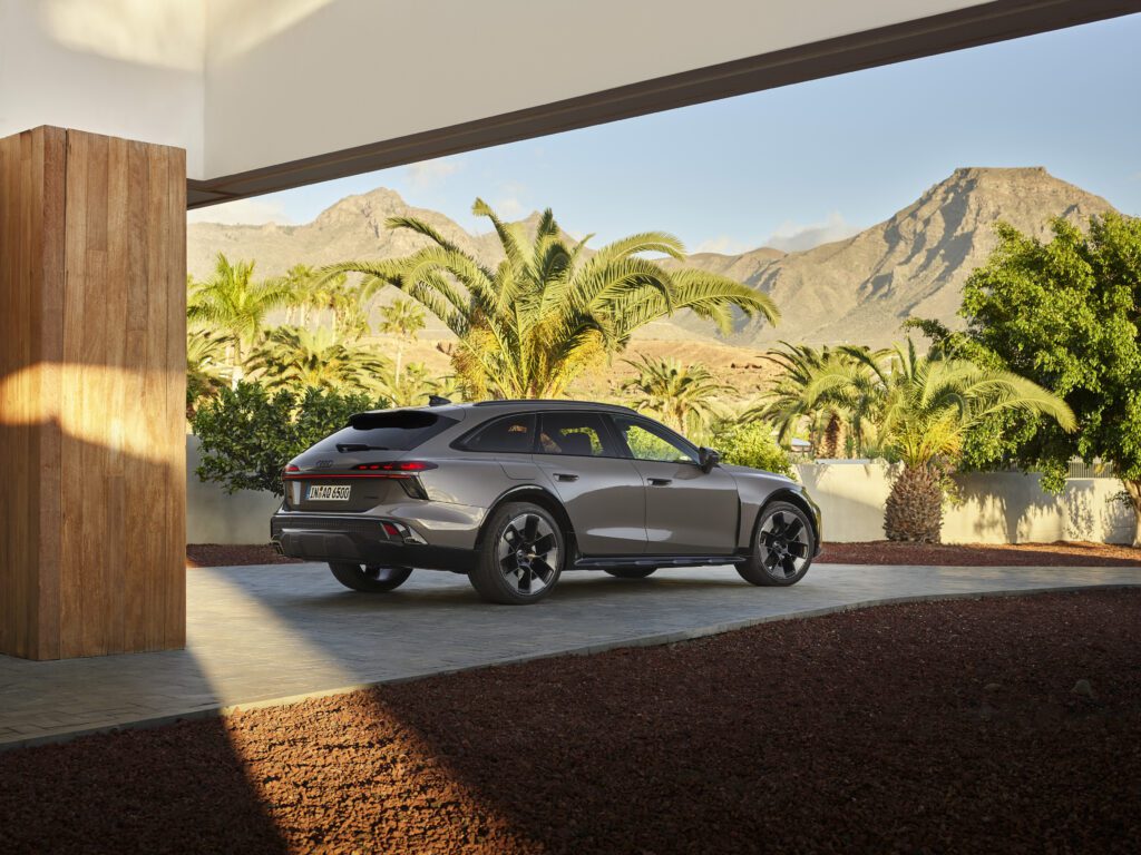 Static three-quarter rear view of the Audi A6 allroad (plateau grey metallic) set in modern architecture with a striking mountain panorama in the background (static shot).